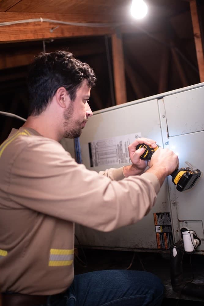 Josiah Hirsh in an attic, driving a screw into the side panel of an air handler with a cordless drill