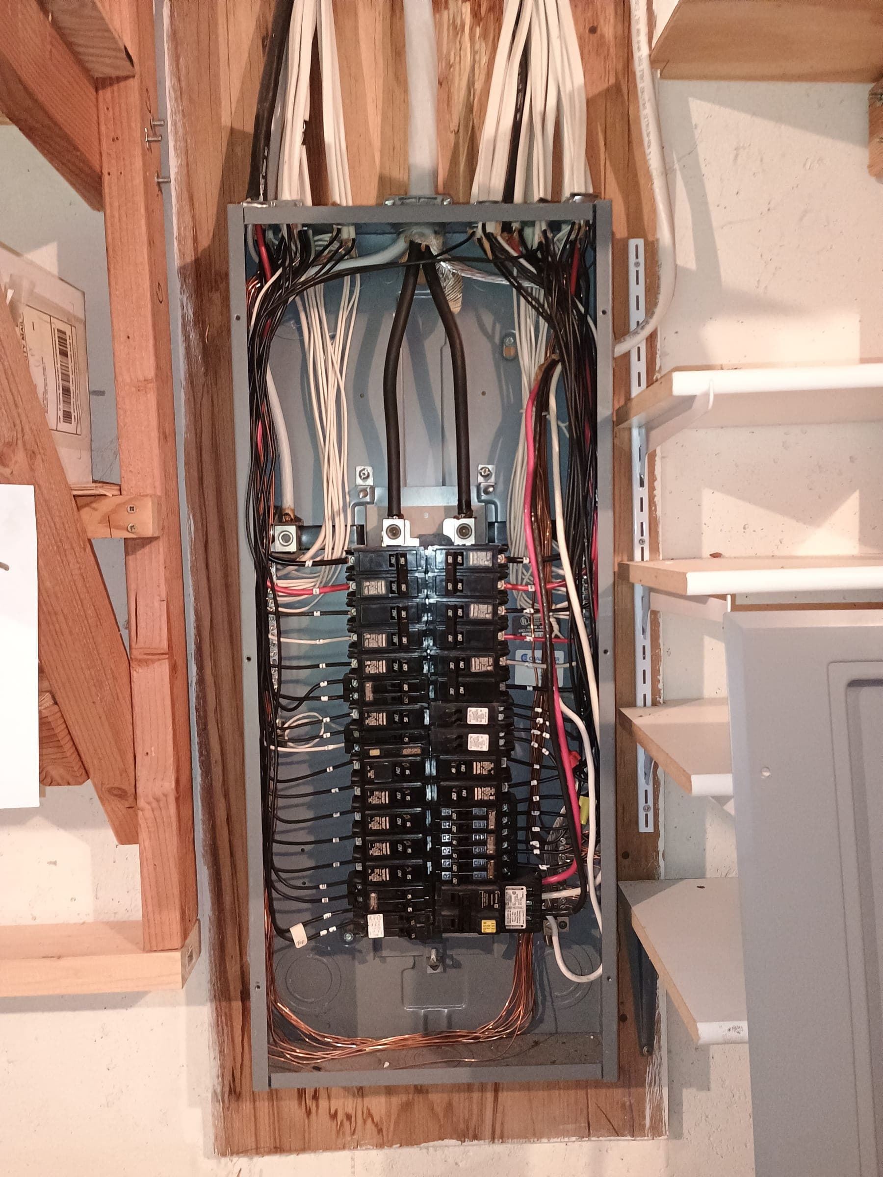 A breaker panel with its cover off between wooden studs, the breakers in two columns and the branch wiring neatly dressed down both sides